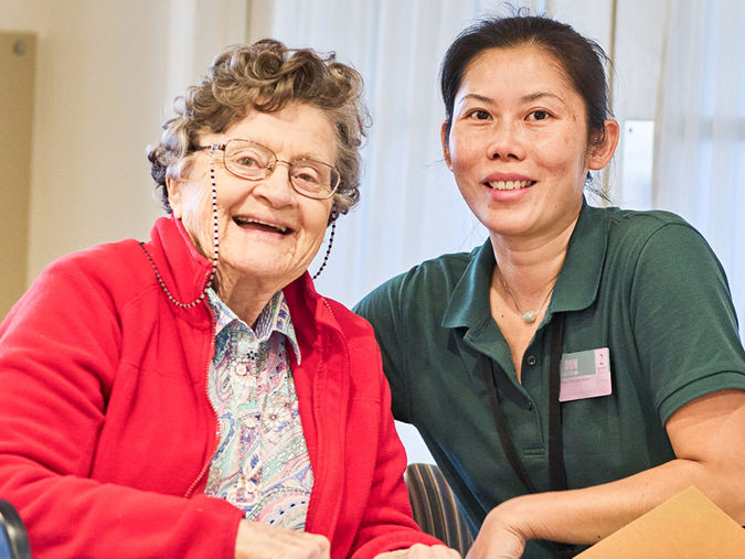Senior woman in a red jacket smiling beside a caregiver in a green shirt, both seated indoors with a light background.