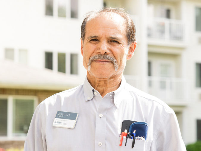 Man with a mustache wearing a name tag and pens in his shirt pocket, standing in front of a building with balconies.