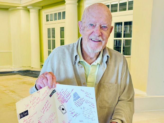 Senior man smiling, holding an open card with handwritten messages, standing indoors near large windows and white pillars.