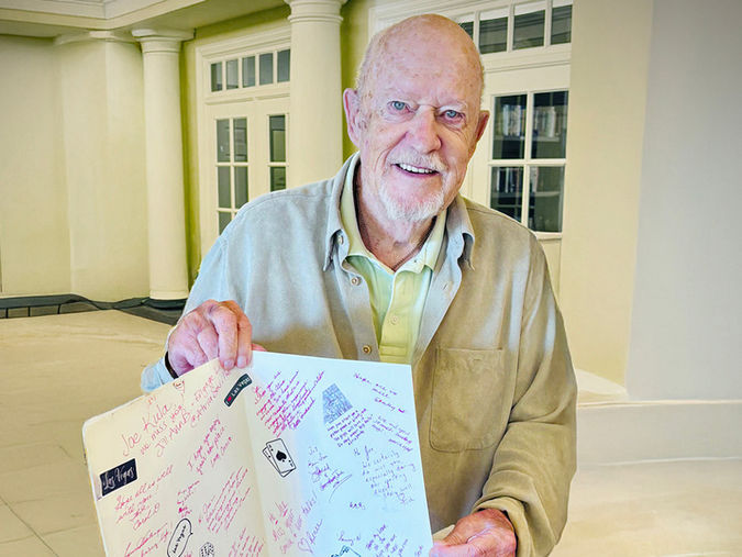 Senior man smiling and holding an open card with colorful handwritten messages, standing in a bright room with white columns.