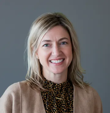 Smiling woman with blond hair wearing a brown sweater and black polka dot blouse against a gray background.