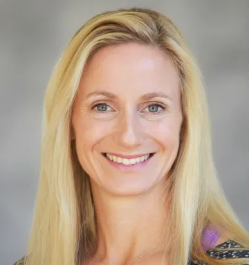 Smiling woman with long blonde hair, wearing a patterned top, against a blurred gray background.