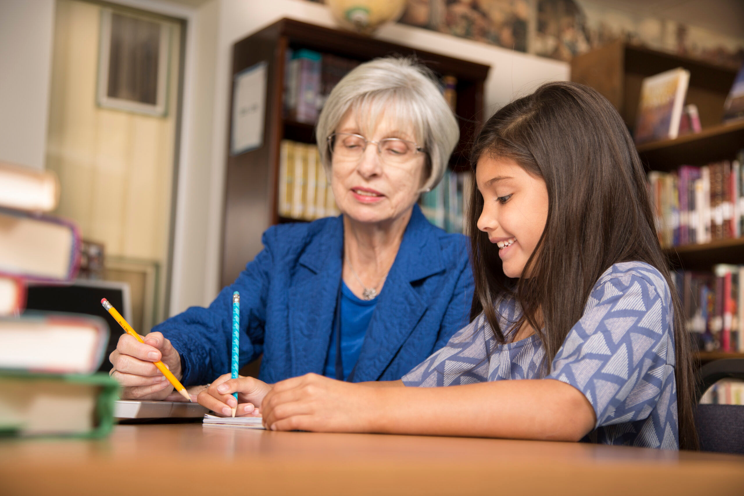 Senior adult teacher, mentor, or tutor assists Latin descent elementary age girl with homework in school classroom or library setting.