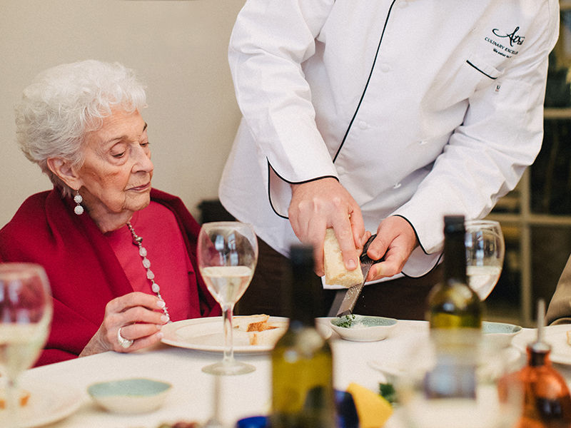 Senior resident dining in the dining room while a server grates fresh parmesan cheese on her meal.