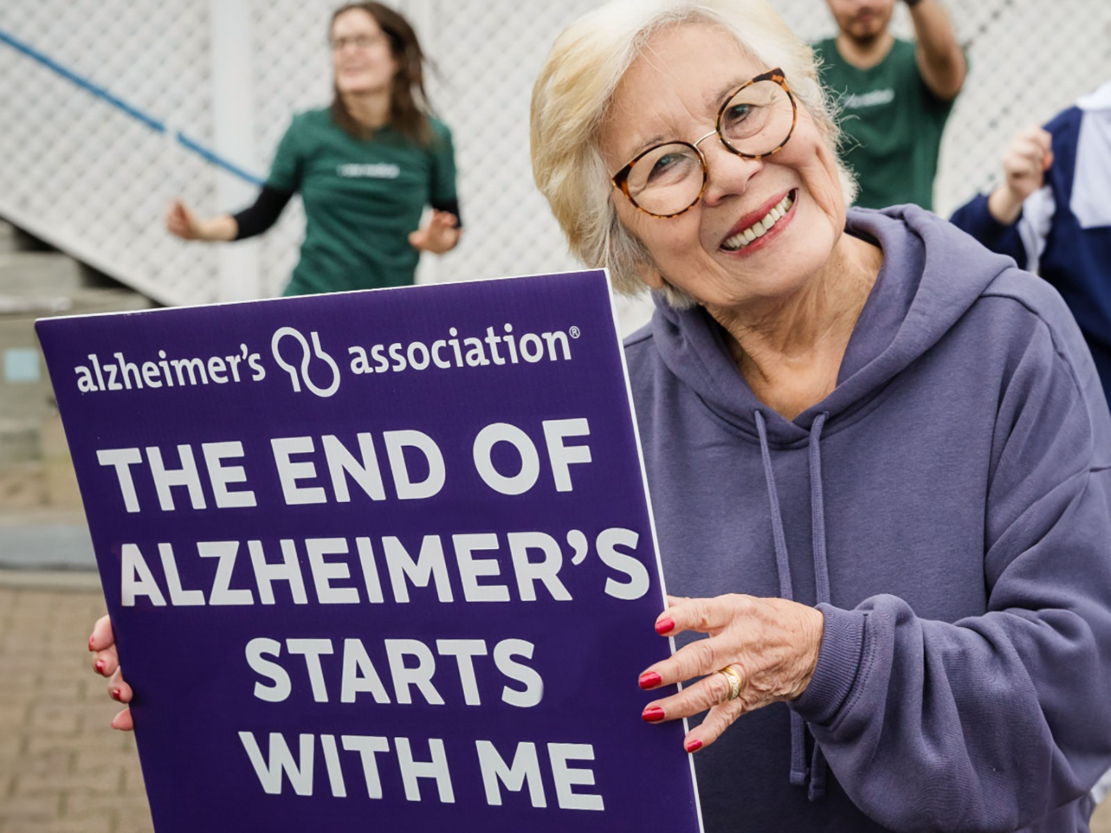 Older woman smiling at the camera and holding a sign that says The End of Alzheimer's Starts with Me