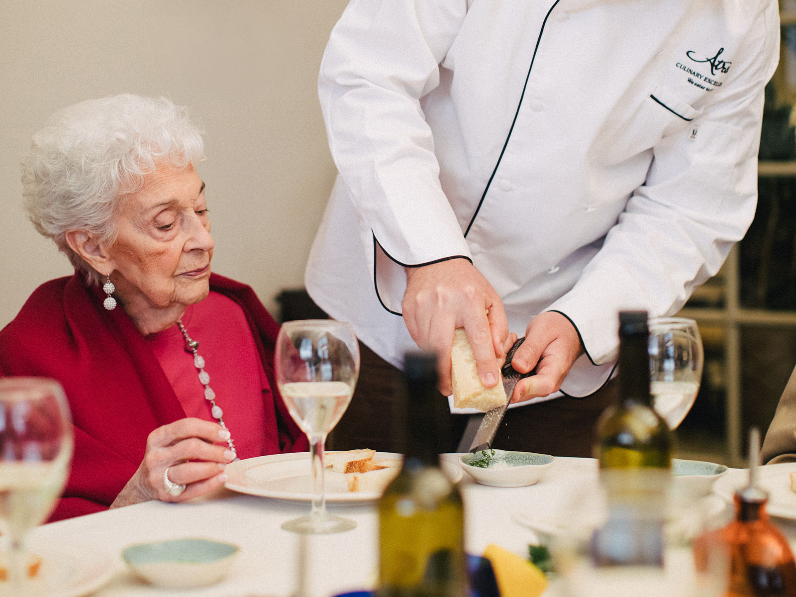 Chef grating Parmesan cheese onto the salad of a discerning older woman wearing red