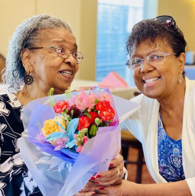 Two older women getting ready to embrace while one holds a bouquet of colorful flowers