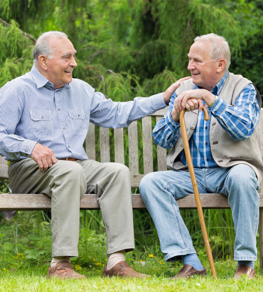 two senior men conversing on a bench with one man having his hand on the other mans shoulder