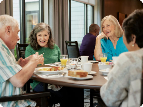 4 senior residents laughing and smiling while enjoying breakfast together in the dining room.