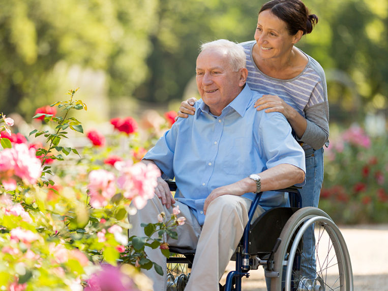 Older woman standing behind man in a wheelchair while they look at flowers outdoors