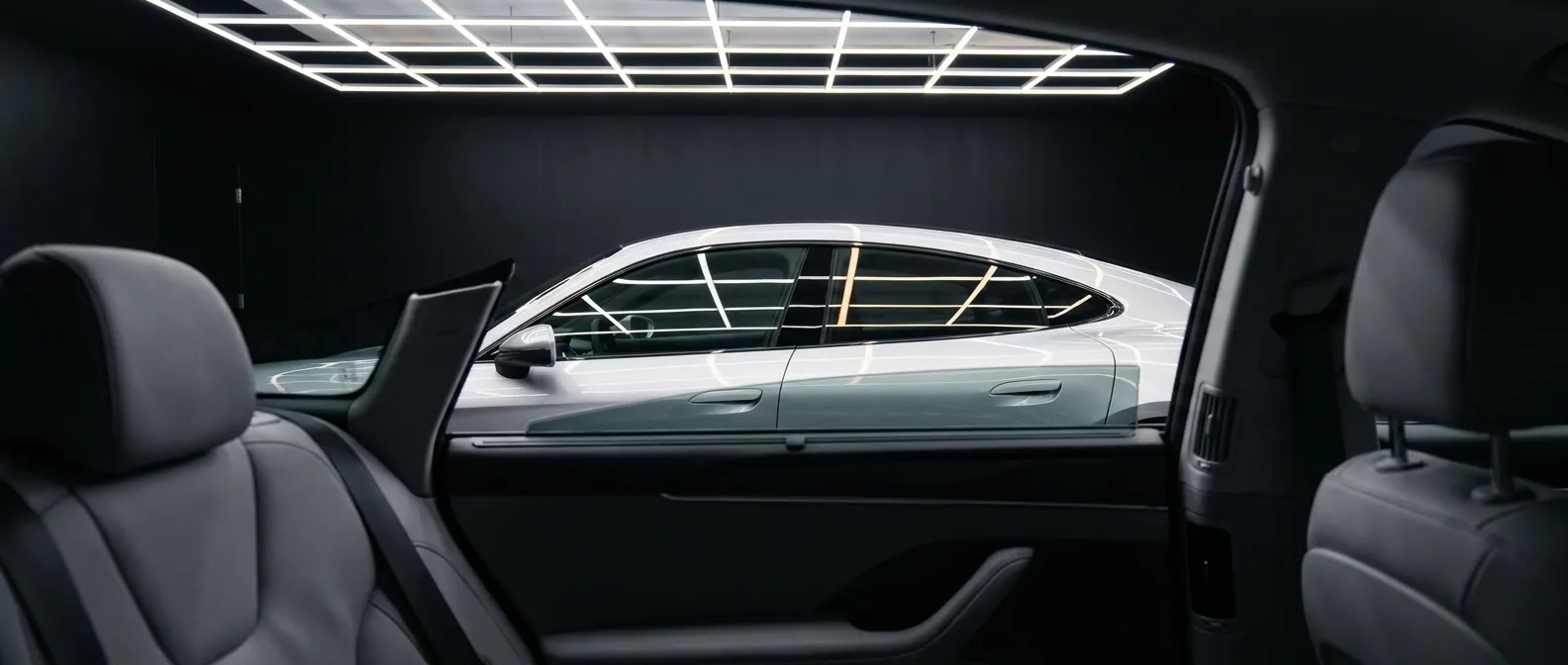 Backseat view of a car interior with gray leather seats and door panel, looking out at a silver sedan parked beside in a dark studio with square grid ceiling lights reflecting on the sedan's windows and glossy body.