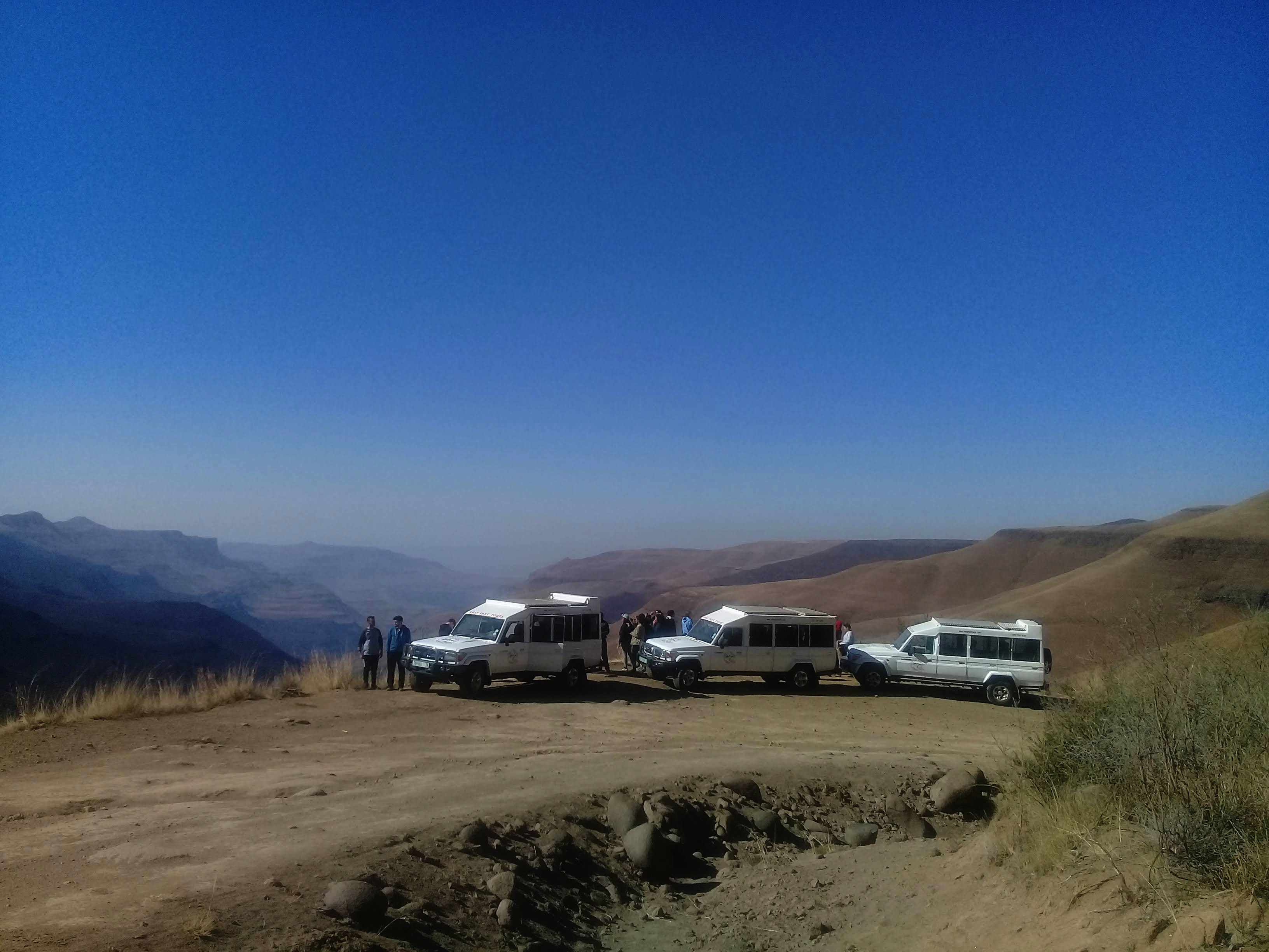 Three specialised 4x4 vehicles are parked on a dirt road, the passengers stand next to the vehicles and gaze out over the dramatic valley to dissappears into the horizon