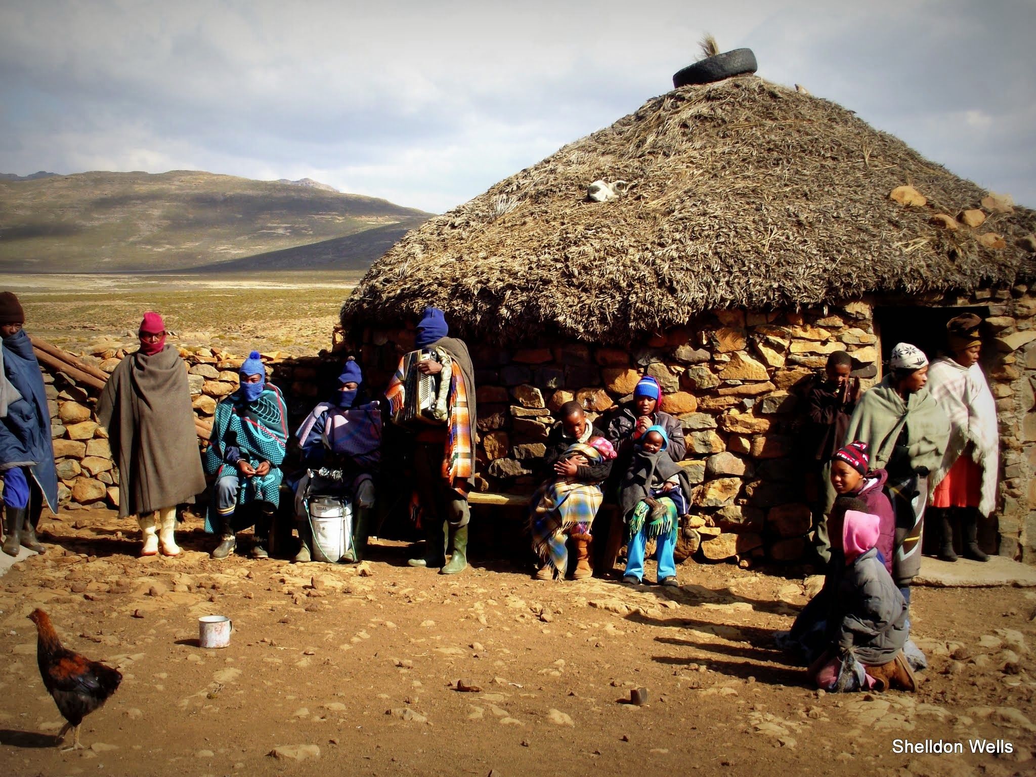A group of Basotho people stand together facing the camera. The people are dressed in traditional blankets. The stand in front of a traditional basotho hut with a grass roof and stone walls.