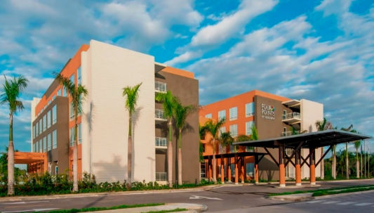 Exterior view of the modern Four Points by Sheraton hotel building on a sunny day, featuring an orange and white facade, a covered entrance, and surrounding palm trees.