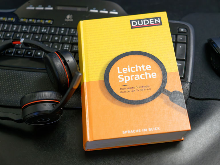 Das Buch zur Leichten Sprache aus dem Duden-Verlag liegt neben einem Headset auf einer Tastatur.