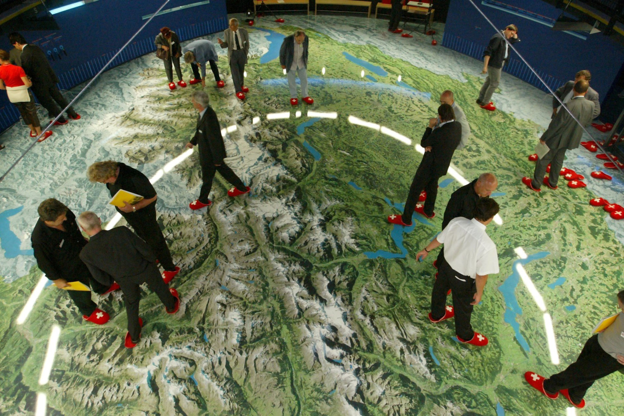 Mehrere Menschen in Pantoffeln stehen auf einem grossen Luftbild der Schweiz.