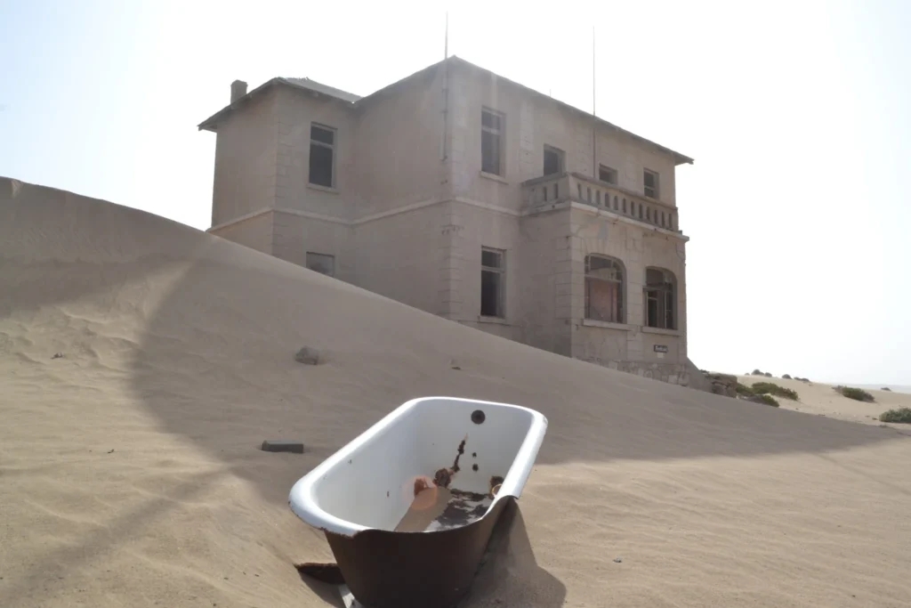 Old bathtub standing in desert sand in front of an abandoned Kolmanskop house