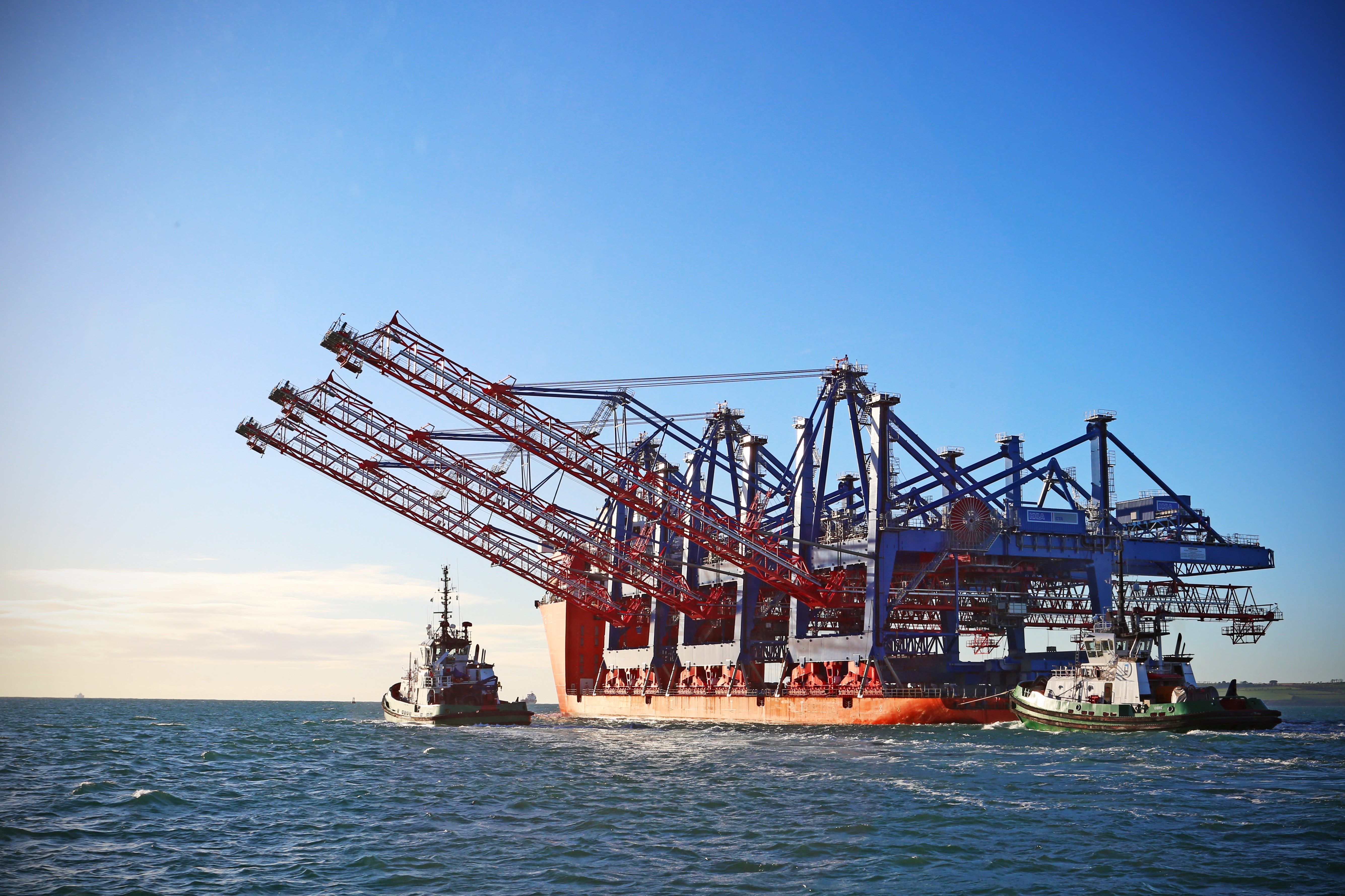 A large cargo ship with cranes is anchored in the ocean, with a tugboat assisting in the foreground under a clear blue sky.