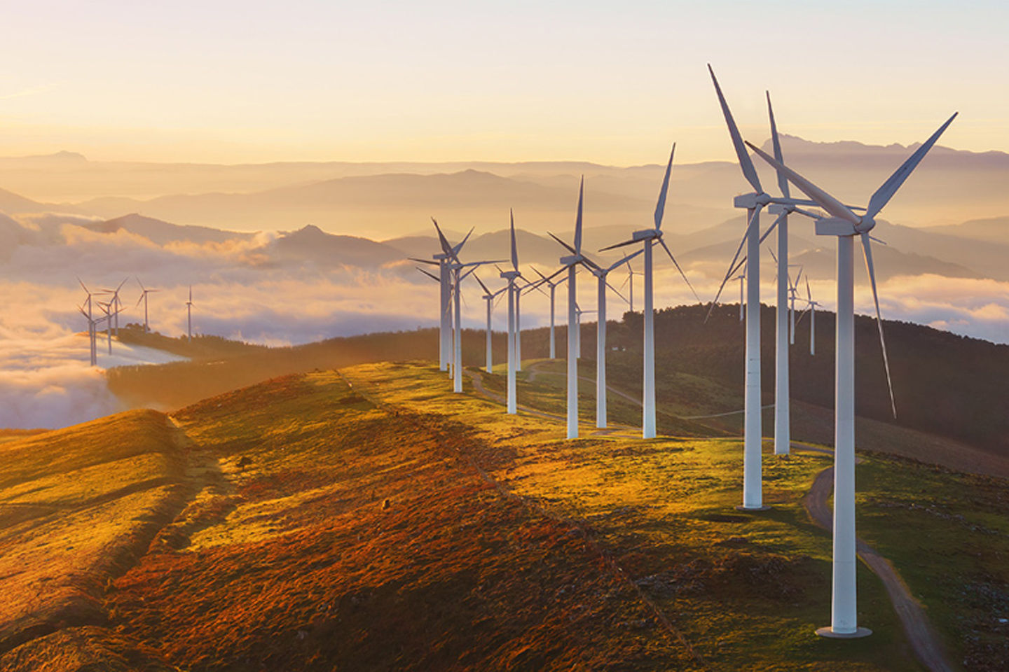 Wind turbines on a hillside at sunrise, surrounded by misty valleys and distant mountains.