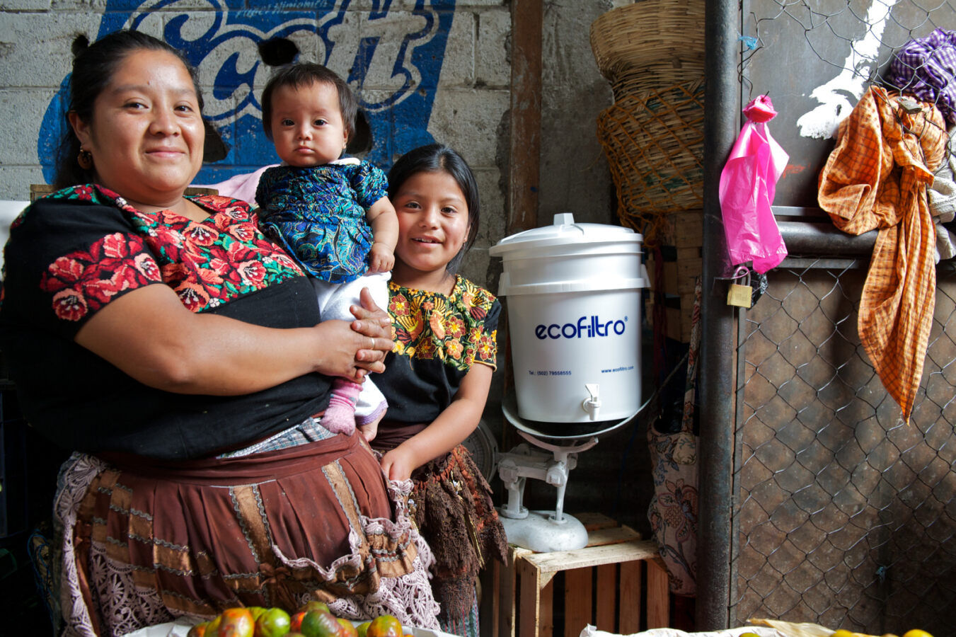 A woman holds a baby with a young girl beside them with their Ecofiltro water filter in Guatemala