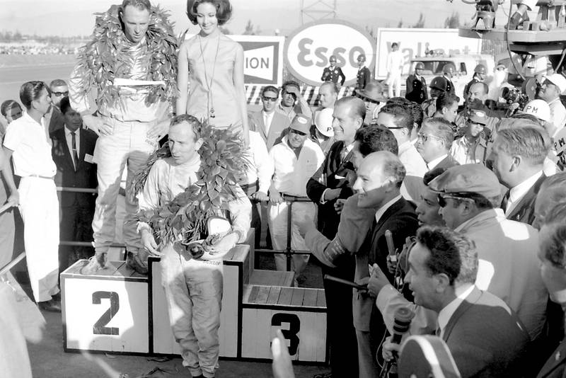 Race winner Dan Gurney stands on the podium with 2nd placed John Surtees standing below. Stirling Moss stands to the right with the media.