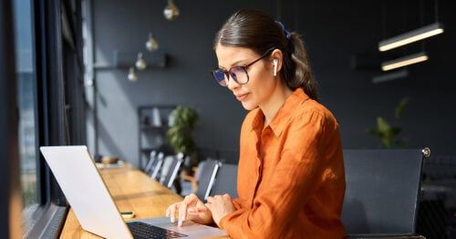 Junge Frau am Laptop mit oranger Bluse, Brillen, in einem Meetingraum, konzentriert beim Arbeiten.