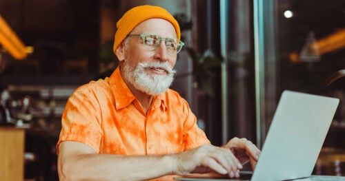 Smiling senior man, bearded hipster wearing yellow hat using laptop computer, working online sitting in modern cafe. Stylish businessman planning startup. Successful business concept